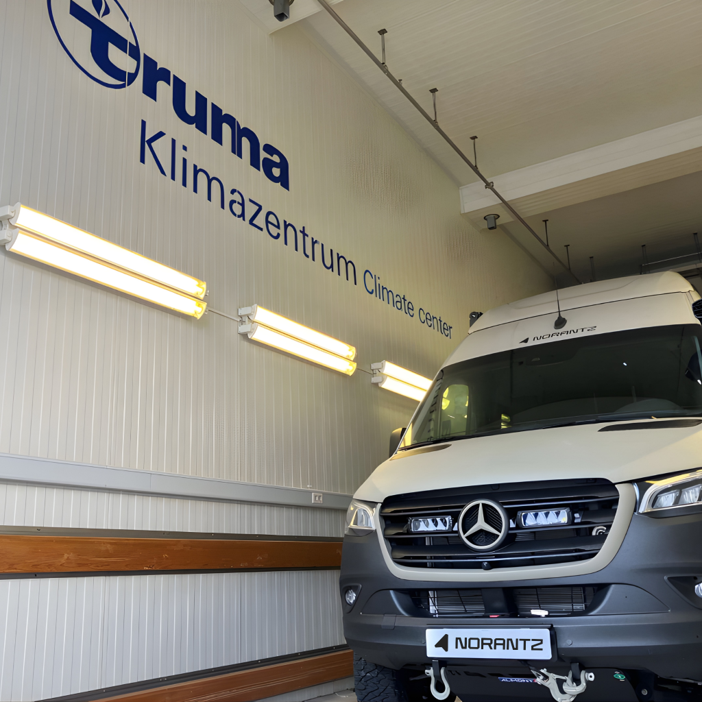 Front view of a Norantz camper inside the Truma climate chamber, with the "Klimazentrum Climate centre" logo on the wall. Vista frontal de una camper Norantz dentro de la cámara climática de Truma, con el logotipo "Klimazentrum Climate center" en la pared.